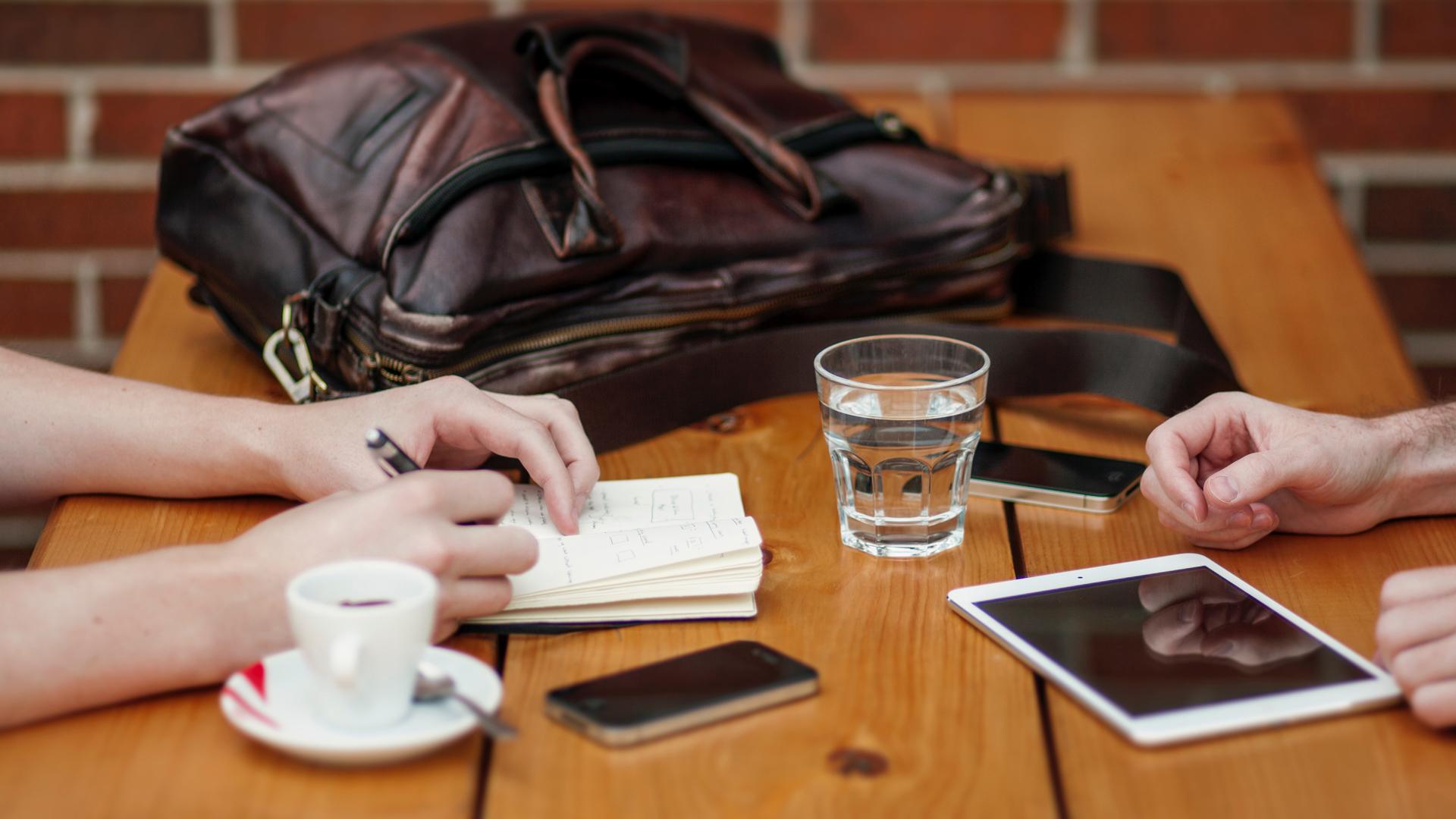 picnic table near brick wall with leather bag, glass of water, notebook, phones and table, with hands on either side