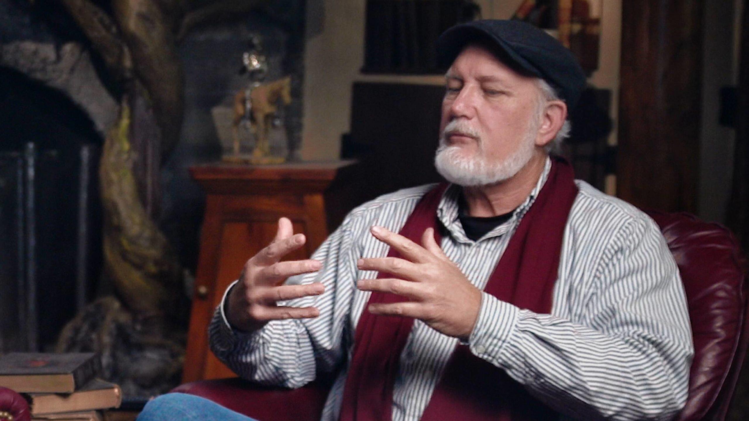 Roy Williams sitting in leather chair, gesturing, wearing red scarf and black cap